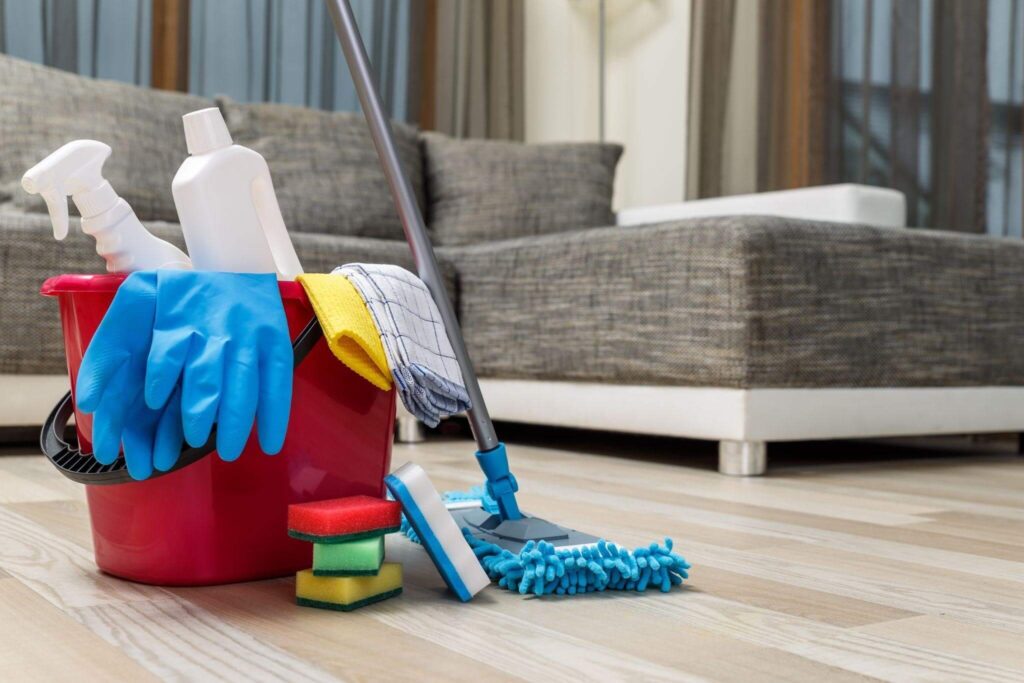 Un seau rouge contenant des produits d'entretien, des éponges, des gants, des vaporisateurs et une serpillière trône sur un parquet, devant un canapé gris, dans un salon — un cadre chaleureux aussi accueillant qu'un jardin en octobre en Provence.