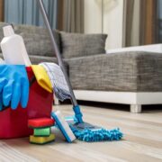 Un seau rouge contenant des produits d'entretien, des éponges, des gants, des vaporisateurs et une serpillière trône sur un parquet, devant un canapé gris, dans un salon — un cadre chaleureux aussi accueillant qu'un jardin en octobre en Provence.