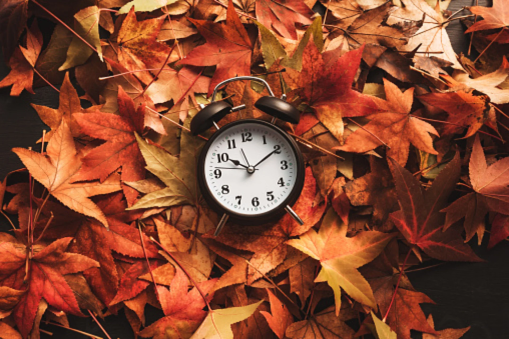 Un réveil noir indiquant 2 h 54 est entouré d’un tas de feuilles d’automne aux nuances d’orange, de rouge et de marron — un rappel saisissant de la date du changement d’heure de 2026.