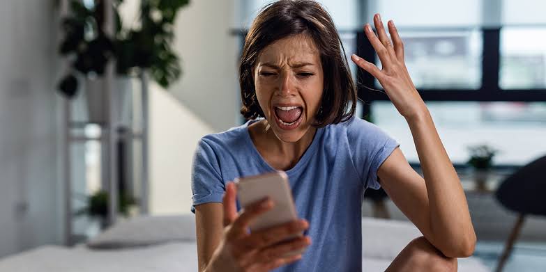 Une femme est assise chez elle, les yeux rivés sur son téléphone, l'air agacé, la bouche grande ouverte comme si elle criait et une main levée en signe d'exaspération — peut-être vient-elle de recevoir un nouvel appel indésirable, ignorant que le démarchage en matière d'adaptation du logement est interdit, ou se demandant si elle devrait signaler un appel abusif à SignalConso.