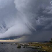 Un nuage d'orage spectaculaire et tourbillonnant se forme au-dessus d'une étendue d'eau près d'un rivage rocheux, avec en arrière-plan un ciel gris foncé et des rayons de soleil qui percent par endroits — à l'image de ces caprices météorologiques qui marquent le moment idéal pour planter « à la Sainte-Catherine » ou commencer un « jardin en novembre » en Provence.
