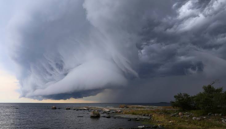 Un nuage d'orage spectaculaire et tourbillonnant se forme au-dessus d'une étendue d'eau près d'un rivage rocheux, avec en arrière-plan un ciel gris foncé et des rayons de soleil qui percent par endroits — à l'image de ces caprices météorologiques qui marquent le moment idéal pour planter « à la Sainte-Catherine » ou commencer un « jardin en novembre » en Provence.
