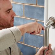 Un homme utilise un tournevis pour fixer une barre d'appui métallique à un mur blanc, à côté de murs carrelés de bleu, une étape importante lorsqu'on cherche à financer l'aménagement de la salle de bains d'un parent ou qu'on envisage des mesures de sécurité pour les pièces où les personnes âgées tombent le plus souvent.