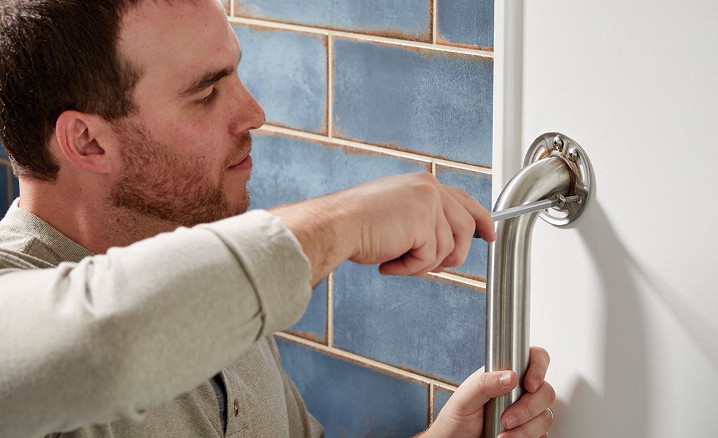 Un homme utilise un tournevis pour fixer une barre d'appui métallique à un mur blanc, à côté de murs carrelés de bleu, une étape importante lorsqu'on cherche à financer l'aménagement de la salle de bains d'un parent ou qu'on envisage des mesures de sécurité pour les pièces où les personnes âgées tombent le plus souvent.
