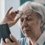 Une femme âgée remonte ses lunettes tout en plissant les yeux pour regarder son smartphone ; elle semble avoir du mal à lire l'écran. Elle présente peut-être des symptômes de la DMLA, ce qui pourrait nécessiter l'utilisation d'outils tels que la grille d'Amsler ou un meilleur éclairage de son logement pour l'aider à voir.