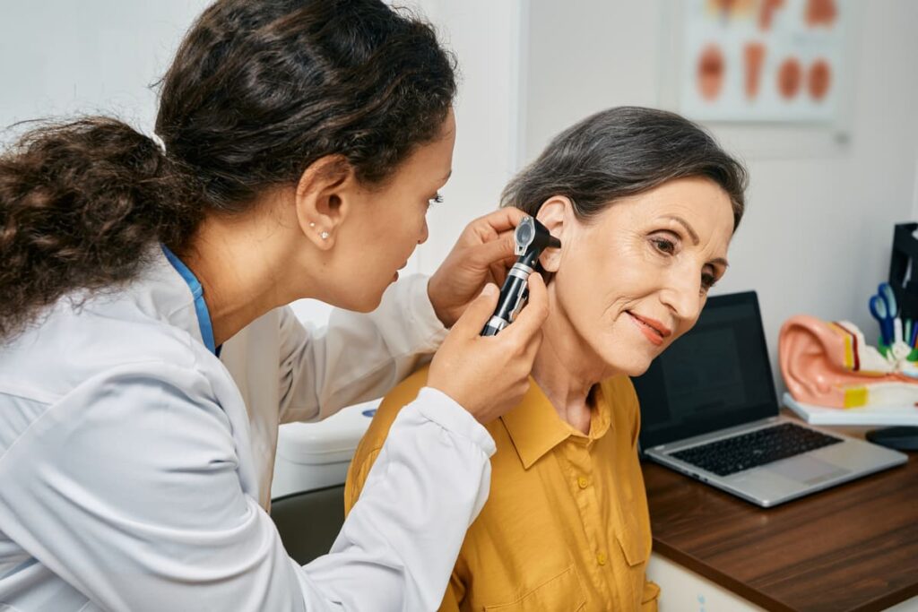 Dans un cabinet médical, un professionnel de santé examine l'oreille d'une femme âgée à l'aide d'un otoscope, avec un ordinateur portable et des planches anatomiques en arrière-plan. Cette prise en charge attentive revêt une importance particulière pour les patients sourds ou malentendants, conformément au décret n° 2026-838, qui garantit l'accessibilité et un accompagnement adapté lors des consultations médicales.