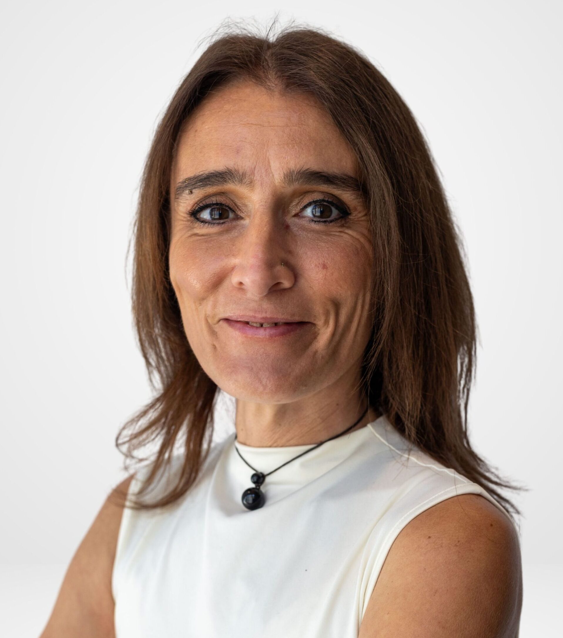 Une aide à domicile d'âge mûr, aux longs cheveux châtains, souriante, les bras croisés, vêtue d'un haut blanc et portant un collier avec un pendentif noir.