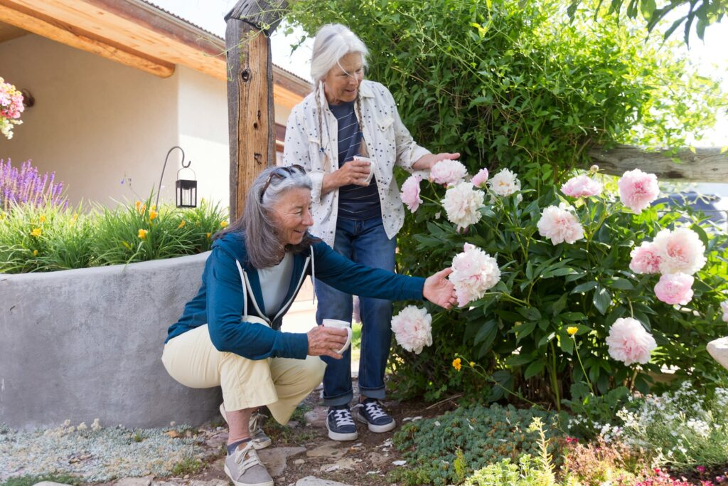 Deux femmes, une tasse à la main, se tiennent dans un jardin et admirent un pivoine en fleurs : une idée de cadeau bien pensée pour une personne âgée vivant de manière autonome.