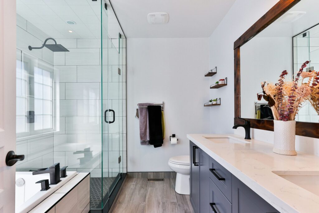 Une salle de bains comprenant une douche vitrée équipée d'une barre d'appui pour prévenir les chutes, un meuble-lavabo gris avec un plan de travail en quartz, des toilettes et un miroir encadré.