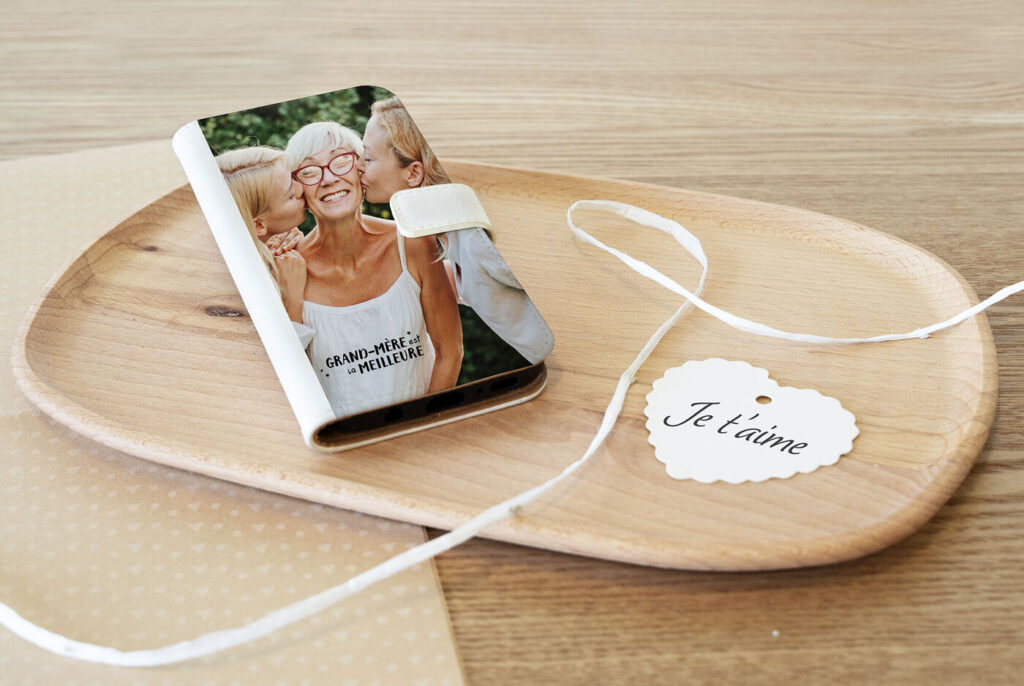 Une coque de smartphone représentant deux femmes embrassant une femme plus âgée sur les joues, avec l'inscription GRAND-MÈRE est la MEILLEURE, posée sur un plateau en bois à côté d'une brochure présentant un fauteuil releveur.