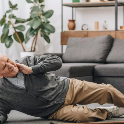 Un homme âgé est allongé sur le sol du salon, souffrant, se tenant la joue, avec sa canne à proximité. Cette scène souligne l'importance de la prévention chutes seniors domicile et du bon aménagement logement seniors chutes.
