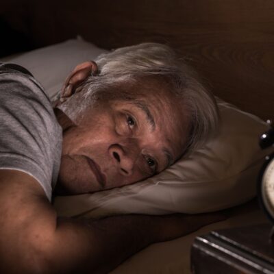 Un homme âgé aux cheveux grisonnants est allongé dans son lit la nuit, l'air inquiet et incapable de dormir. Il est face à un réveil analogique qui indique 3h55, ce qui reflète les difficultés courantes du sommeil senior et la nécessité d'une aide à domicile ou d'alternatives sûres comme la mélatonine senior.