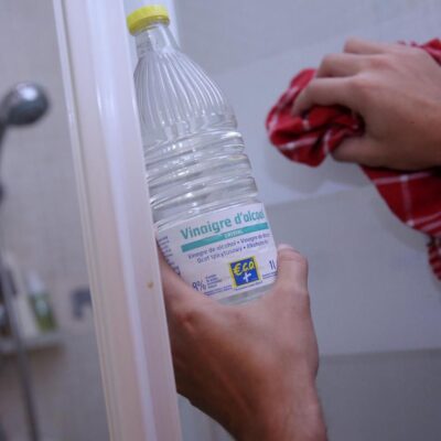 Une personne nettoie une porte de douche avec un chiffon rouge dans une main et une bouteille de vinaigre blanc salle de bain dans l'autre, soulignant son efficacité pour un entretien impeccable de la salle de bain.