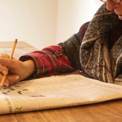 Une personne portant des lunettes et une chemise à carreaux est assise à une table en bois, concentrée à remplir une grille de mots croisés dans un journal, peut-être en train de faire une pause après avoir été aide ménagère du CESU.