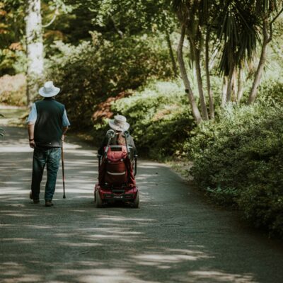 Un homme âgé avec une canne marche à côté d'une femme dans un scooter motorisé le long d'un chemin ombragé et bordé d'arbres dans un parc, tous deux portant des chapeaux de couleur claire. Cette scène reflète l'accompagnement handicap moteur cognitif domicile et le label Cap'Handéo prestataire handicap.