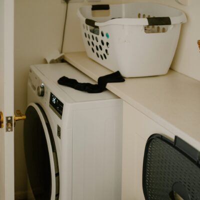 Une buanderie avec une machine à laver à chargement frontal, un panier à linge blanc sur le comptoir, un panier en osier et une chaussette noire drapée sur le bord du comptoir. La porte de la pièce est partiellement ouverte.