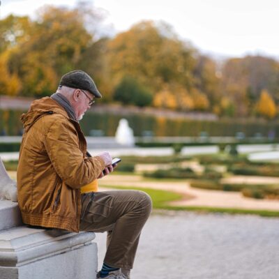 Un homme d'un certain âge, coiffé d'une casquette plate et vêtu d'une veste marron, est assis sur un banc en pierre dans un parc près d'arbres d'automne, de jardins et de fontaines. Il consulte son téléphone, peut-être à la recherche d'une aide à domicile fiable Arles. Un sac à dos est posé à côté de lui.