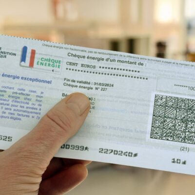 Une main tient un « Chèque Énergie » français d'une valeur de 100 euros, destiné à aider au paiement des factures d'énergie et soumis aux conditions d'utilisation du Chèque Énergie. Le chèque comporte les logos officiels, un code QR et les modalités d'utilisation. L'arrière-plan est flou, ce qui suggère un décor d'intérieur.