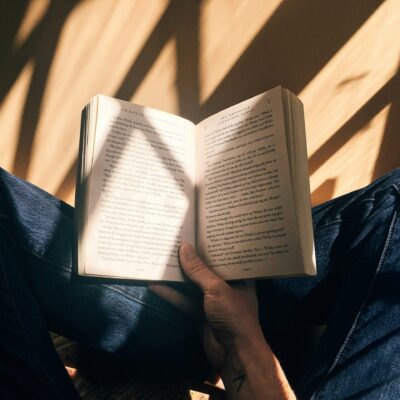 Une personne portant un jean bleu est assise les jambes croisées, tenant un livre ouvert sur ses genoux. La lumière du soleil et les ombres tombent sur les pages du livre et sur ses jambes.