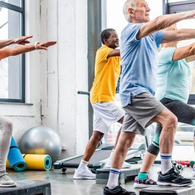 Un groupe de personnes âgées, hommes et femmes, s'exerce sur des plates-formes de marche dans un studio de gymnastique lumineux, levant les bras parallèlement au sol, tout en pratiquant ensemble une activité physique adaptée aux seniors.