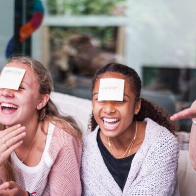 Quatre personnes d'âges différents sont assises ensemble, riant, chacune avec une note autocollante sur le front alors qu'elles s'amusent à jouer à des jeux maison sans accessoires. Une femme fait des gestes animés, ce qui ajoute à la vivacité et à la joie de la scène.
