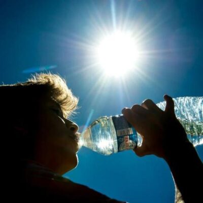 Une personne pratique l'hydratation senior chaleur, en buvant dans une bouteille d'eau sous un soleil radieux, dans un ciel bleu clair, la lumière du soleil créant un effet de brillance autour de sa silhouette.