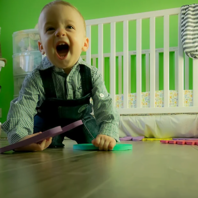 Un bambin souriant portant des bretelles rampe sur un plancher en bois, tenant des pièces de puzzle en mousse colorées, dans une chambre d'enfant lumineuse avec un berceau blanc et des murs verts, surveillée de près par une aide à domicile attentive.