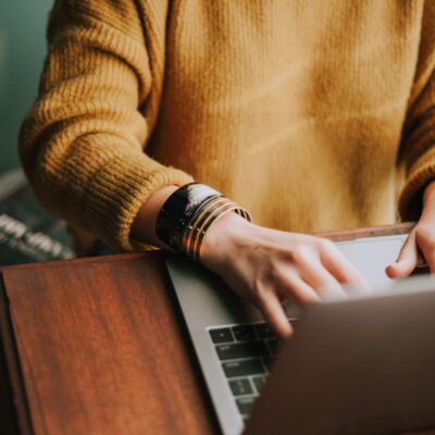 Une personne portant un pull jaune et des bracelets tape sur un ordinateur portable à un bureau en bois, cherchant peut-être un emploi aide à domicile week-end, avec un livre partiellement visible à proximité.