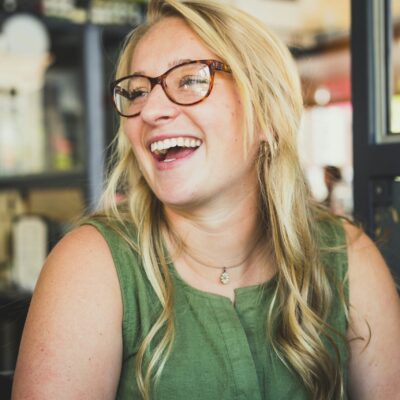 Une femme aux longs cheveux blonds et aux lunettes sourit et rit, assise à l'intérieur. Elle porte un haut vert sans manches et un collier, représentant la stabilité équipes aide à domicile avec la lumière du soleil qui entre par une fenêtre voisine.