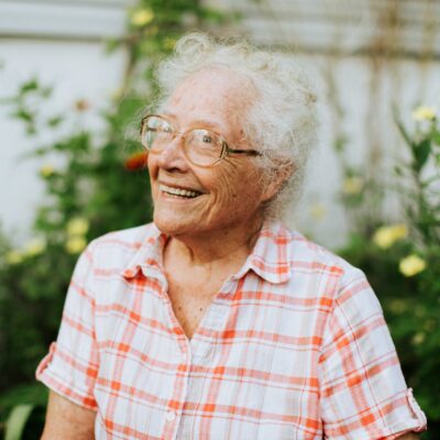 Une femme âgée aux cheveux blancs bouclés et aux lunettes sourit, vêtue d'une chemise à carreaux rouges et blancs, debout dans un jardin avec des plantes vertes et des fleurs jaunes en arrière-plan.