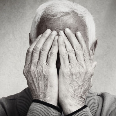 Une personne âgée aux cheveux blancs se couvre le visage des deux mains, ne laissant apparaître que ses mains et son front. Cette image en noir et blanc met en évidence les rides et la texture, capturant les signes dépression senior à travers une émotion subtile.