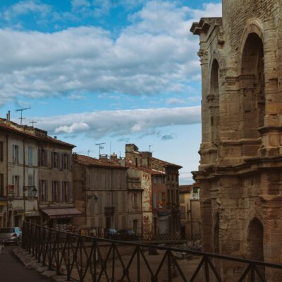 Une rue européenne étroite d'Arles bordée de bâtiments anciens, dont une grande structure en pierre avec des arches sur la droite. Des personnes, des voitures et une aide à domicile seniors Arles sont visibles sous un ciel partiellement nuageux avec des taches de bleu.