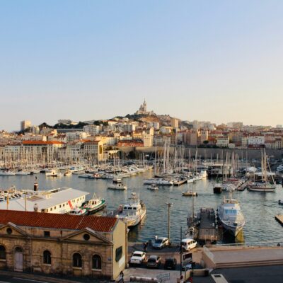Un port rempli de nombreux voiliers et yachts, bordé de bâtiments historiques aux toits de tuiles rouges. En arrière-plan, une ville s'élève sur une colline surmontée d'une basilique - une vue emblématique de Marseille, où le service à la personne Marseille prospère.