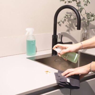 Une personne vaporise de la solution de nettoyage sur un comptoir près d'un évier, tenant un chiffon sombre pour essuyer les pièces de monnaie, montrant le soin méticuleux du ménage à domicile Nîmes. Un flacon pulvérisateur et un vase avec de la verdure se trouvent à l'arrière-plan.
