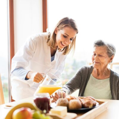 Une aide-soignante souriante en blouse blanche verse du jus d'orange à une femme âgée assise à une table garnie d'aliments pour le petit-déjeuner tels que des croissants, des fruits et de la confiture, prête à partager des astuces telles que la manière de nettoyer un Thermomix.