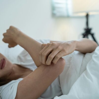 Une femme est allongée dans son lit, semblant mal à l'aise alors qu'elle tient son coude d'une main, peut-être en raison d'une douleur ou d'une gêne causée par des punaises de lit. L'arrière-plan doucement éclairé comporte une lampe et une literie de couleur claire.