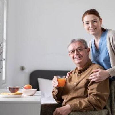 Un homme âgé est assis à une table, devant un jus de fruits et des fruits, et sourit. Une jeune femme, sans doute une aide-soignante, se tient derrière lui, les mains posées sur ses épaules, et sourit à la caméra. Leur bonheur reflète peut-être la sérénité que procure la participation à la campagne de vaccination contre la grippe et la Covid-19, qui garantit à tous deux une protection pendant cette saison.
