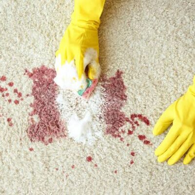 Deux mains gantées de jaune nettoient une tache de liquide rouge sur un tapis clair à l'aide d'une éponge et d'une mousse nettoyante, illustrant ainsi comment le maintien de la propreté peut constituer une alternative efficace à l'EHPAD pour les personnes âgées qui préfèrent rester chez elles.