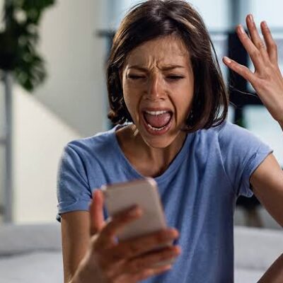 Une femme est assise chez elle, les yeux rivés sur son téléphone, l'air agacé, la bouche grande ouverte comme si elle criait et une main levée en signe d'exaspération — peut-être vient-elle de recevoir un nouvel appel indésirable, ignorant que le démarchage en matière d'adaptation du logement est interdit, ou se demandant si elle devrait signaler un appel abusif à SignalConso.
