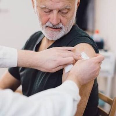 Un professionnel de santé essuie le haut du bras d'un homme âgé, assis et vêtu d'une chemise à manches courtes, afin de le préparer à une injection ou à une vaccination dans un établissement médical. Cette scène souligne l'importance de se faire vacciner lorsqu'on ne peut pas se déplacer, afin de garantir que même les personnes à mobilité réduite aient accès aux vaccinations essentielles, telles que la vaccination contre la grippe et la Covid.