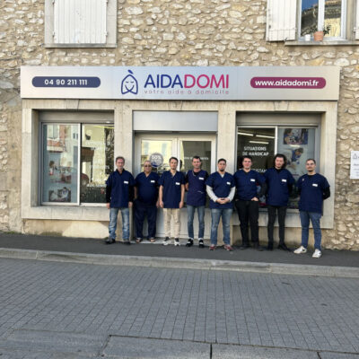 Neuf personnes portant des uniformes bleus font la queue devant un bâtiment en pierre sur lequel on peut lire AIDADOMI, votre aide à domicile. Le numéro de téléphone et le site Internet de l'entreprise sont également visibles au-dessus de l'entrée.
