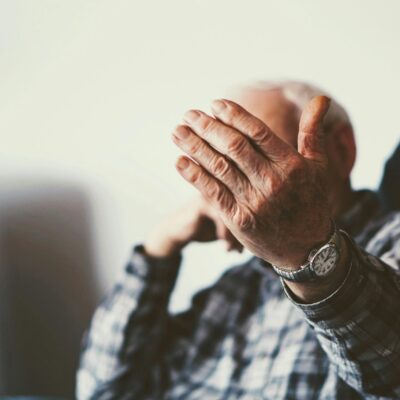 Une personne âgée assise sur une chaise, portant une chemise à carreaux et une montre-bracelet, tend le bras vers l'appareil photo et masque la majeure partie de son visage - un moment qui reflète l'importance de l'aide à domicile Alzheimer maintien domicile. L'arrière-plan est légèrement flou.