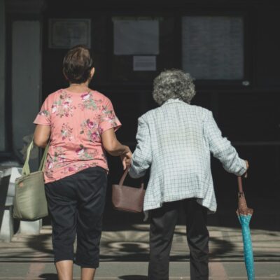 Deux femmes se dirigent main dans la main vers l'entrée d'un immeuble - l'une porte une chemise rose à fleurs et un sac vert, l'autre, aux cheveux gris, s'appuie sur une canne et tient un parapluie bleu - illustrant choisir aide à domicile Arles.