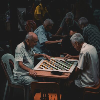 Deux hommes âgés sont assis l'un en face de l'autre à une petite table, jouant intensément à un jeu avec un plateau à carreaux, tandis que d'autres hommes - certains assistés par une aide ménagère du CESU - se rassemblent et jouent à des jeux dans l'arrière-plan faiblement éclairé.