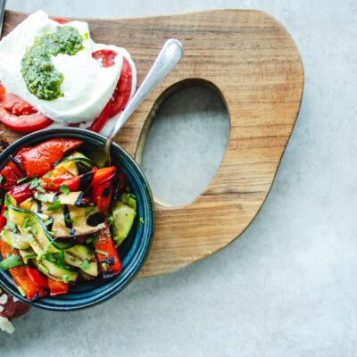 Une planche en bois présente un bol de légumes grillés, une salade fraîche avec des légumes verts et des noix, et des tranches de tomates garnies de mozzarella et de pesto. Des tranches de saumon se trouvent à proximité. L'arrière-plan est une surface de pierre grise.