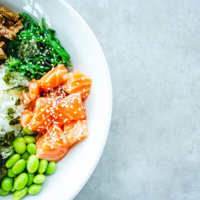 Un bol blanc rempli de riz, de tranches de saumon cru garnies de graines de sésame, d'edamame, d'algues, d'avocat, de légumes verts et de tempeh est posé sur une surface gris clair - parfait pour un repas sain après avoir organisé les services d'aide ménagère du CESU.