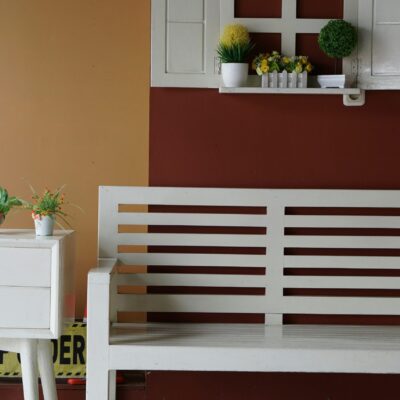 Un banc en bois blanc se trouve à côté d'une petite table blanche avec des plantes en pot. Au-dessus, une fausse fenêtre avec d'autres plantes décore un mur bicolore brun et rouge. Un panneau "Attention" est partiellement visible derrière la table.