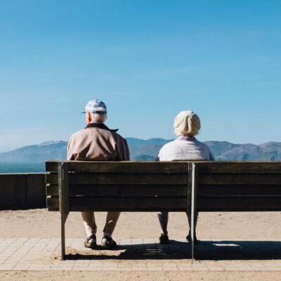Deux personnes âgées sont assises côte à côte sur un banc, profitant d'une vue panoramique sur la montagne et l'eau sous un ciel bleu clair. Ce moment de calme reflète la valeur de Ressources d'Aide pour Personnes Âgées en Ville, qui soutient la vie urbaine paisible des personnes âgées.