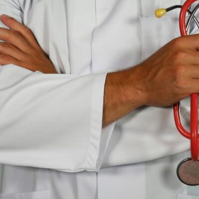 Une personne en blouse blanche se tient debout, les bras croisés, un stéthoscope rouge dans une main, prête à coordonner l'APA urgence retour hospitalisation. Le visage n'est pas visible.