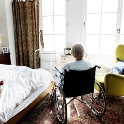 Une personne âgée aux cheveux gris courts est assise dans un fauteuil roulant, face aux grandes fenêtres d'une chambre douillette baignée de lumière naturelle. Elle réfléchit tranquillement au récent refus d'aide senior et à ses sentiments concernant l'aide à domicile imposée.